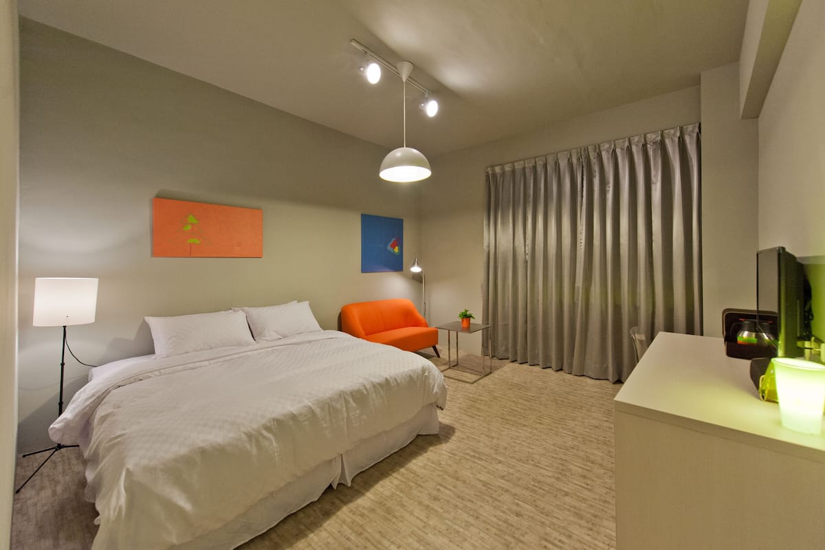 Une chambre d’hôtel avec un grand lit, un fauteuil orange, un bureau avec une lampe et un rideau.