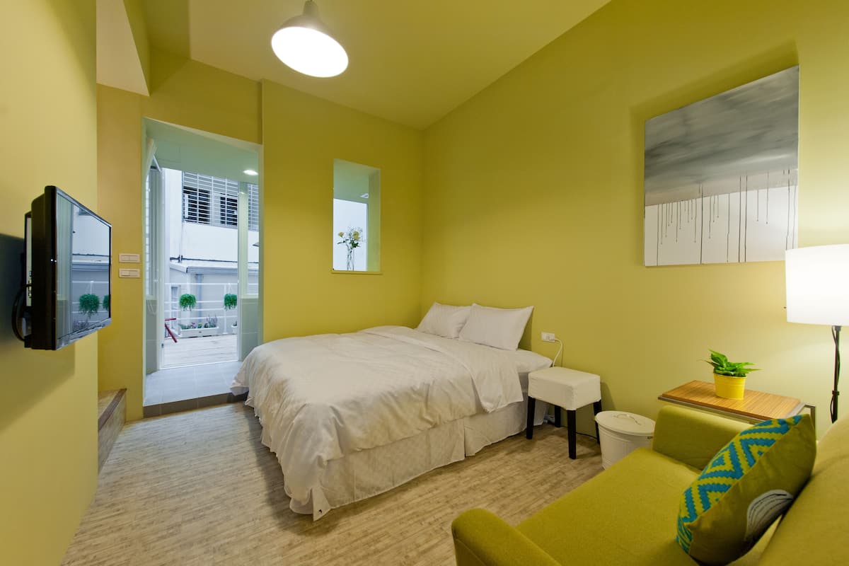 Une chambre d’hôtel avec un lit, une petite table avec une lampe, un fauteuil vert, une télévision et une fenêtre donnant sur l’extérieur.