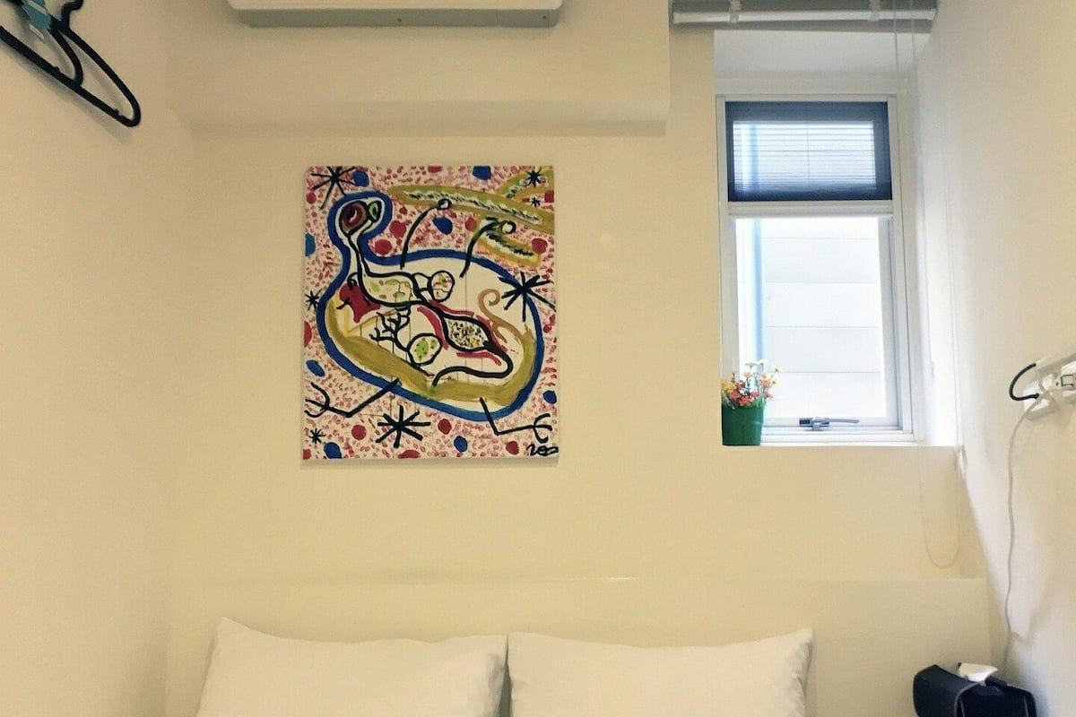 Une chambre à coucher avec un lit, un climatiseur, une fenêtre et un tableau abstrait aux couleurs vives.