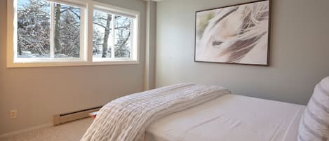 Een slaapkamer met een groot raam, een bed met wit beddengoed en een abstract schilderij aan de muur.