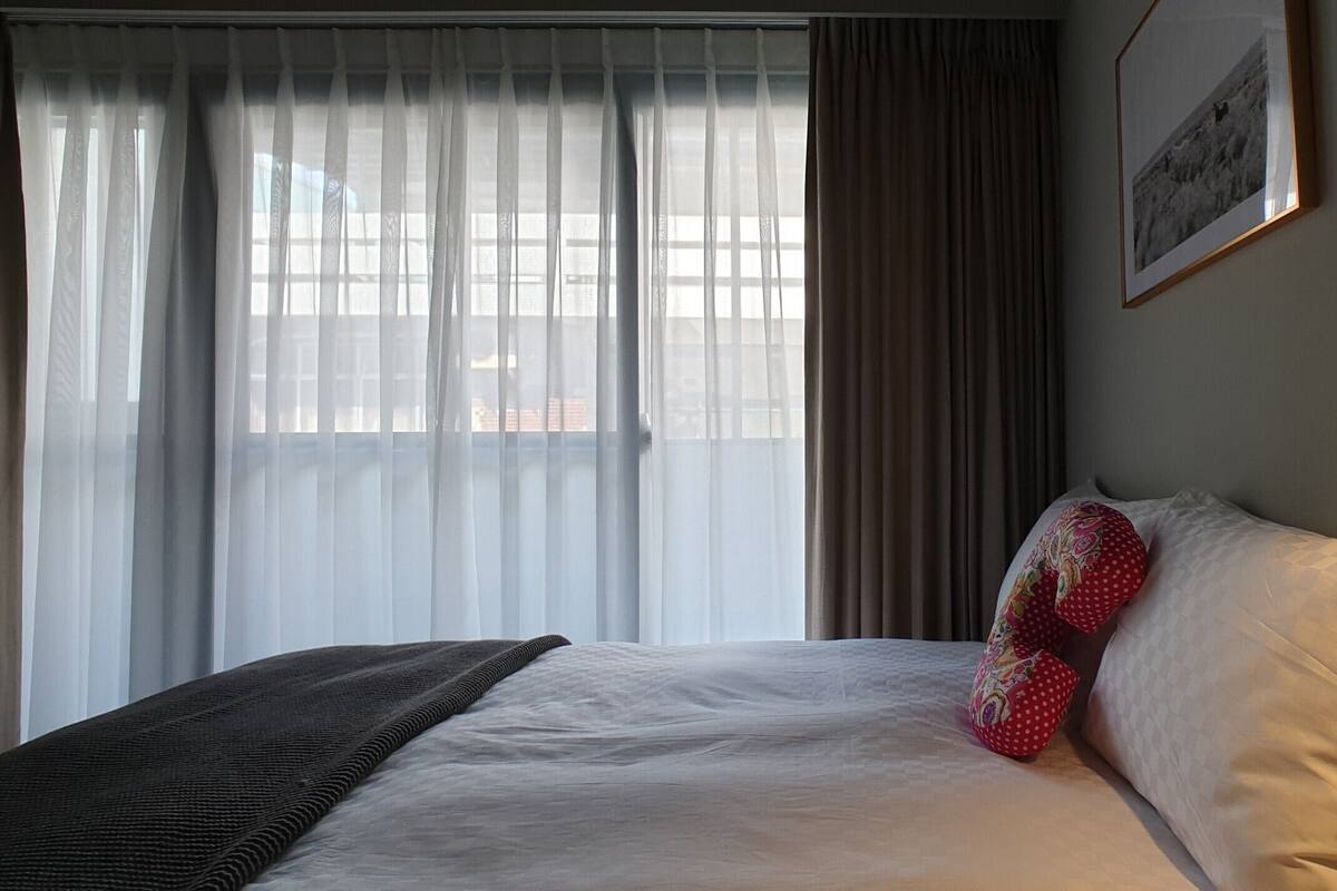 A hotel room with a bed, a window with sheer curtains, a dark-colored throw blanket, a red pillow with a floral pattern, and a framed picture on the wall.