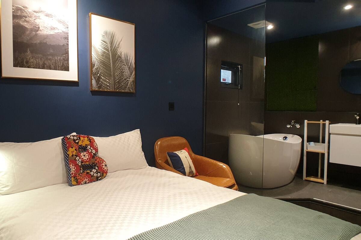 A modern hotel room with a large bed, a chair, a bathtub, and a small bathroom.