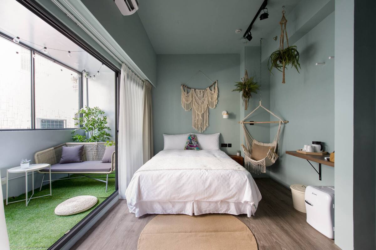 A bedroom with a bed, a hanging chair, a small table, a potted plant, and a window with curtains.