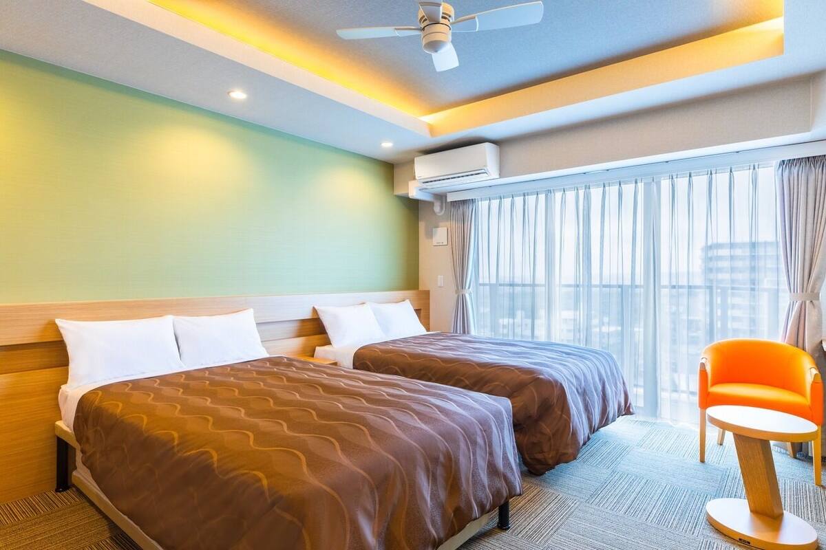 Habitación de hotel con dos camas, ventilador de techo, equipo de aire acondicionado, ventana con cortinas y una pequeña silla naranja.