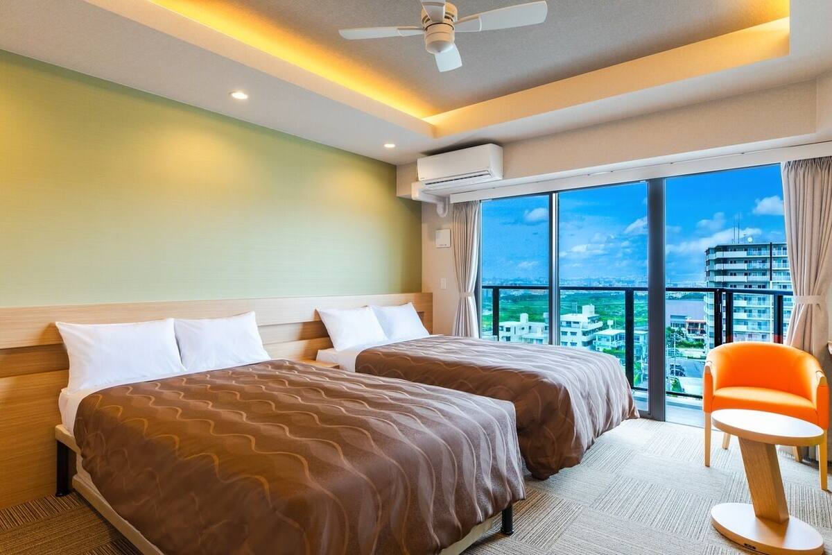 Habitación de hotel con dos camas, una silla, equipo de aire acondicionado, ventilador de techo y una ventana grande con vista a la ciudad.