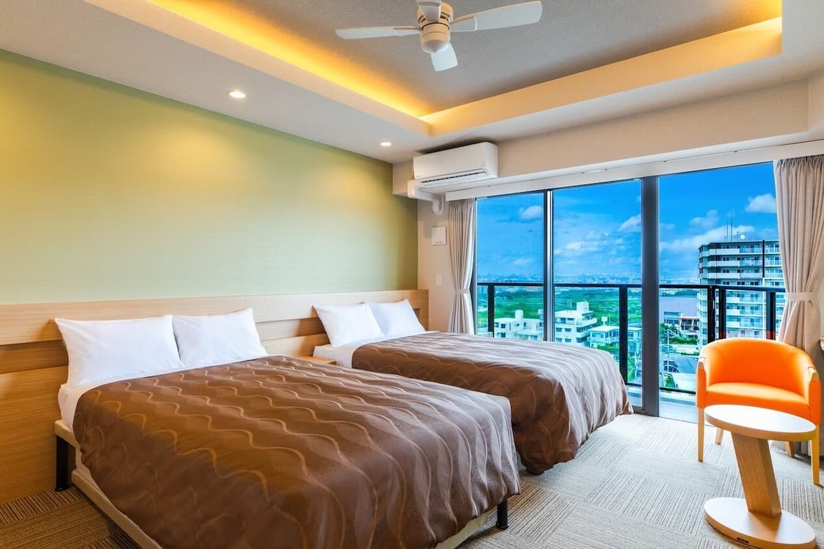 Habitación de hotel con dos camas, una silla, equipo de aire acondicionado, ventilador de techo y una ventana grande con vista a la ciudad.