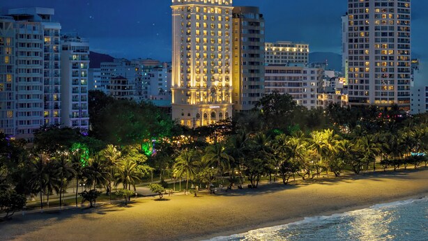 Un hôtel de grande hauteur, doté d’une architecture classique, situé au bord d’une plage bordée de palmiers et offrant une vue sur l’océan.