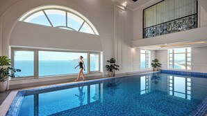 Une grande piscine intérieure avec une vue dégagée sur l’océan, de grandes fenêtres et des plantes en pot.