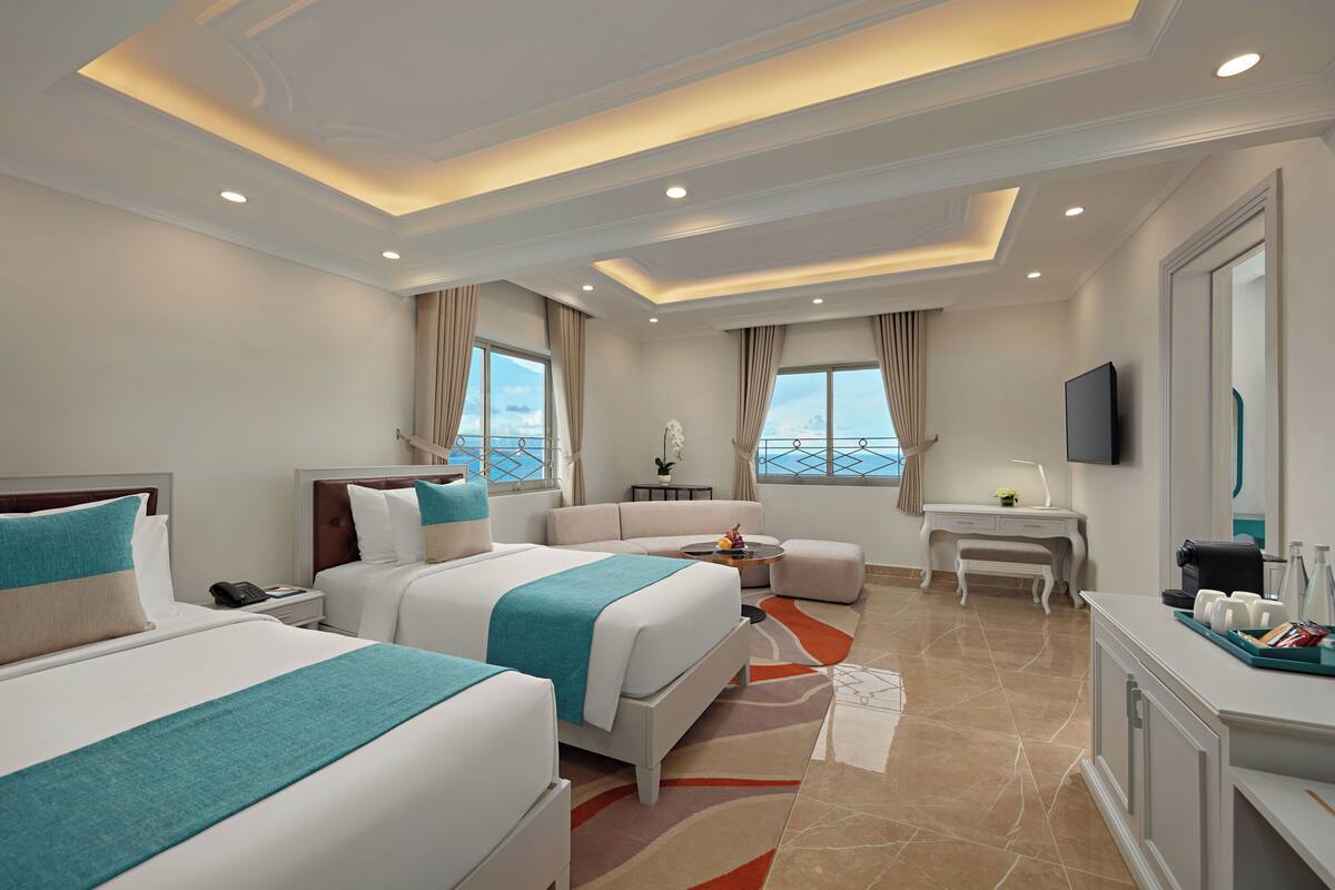 A hotel room with two beds, a sofa, a desk, a TV, and a view of the sea.