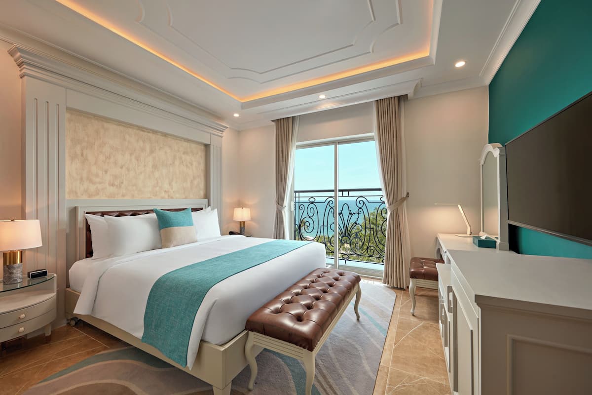 A hotel room with a large bed, a bench, a vanity, and a balcony with a view.