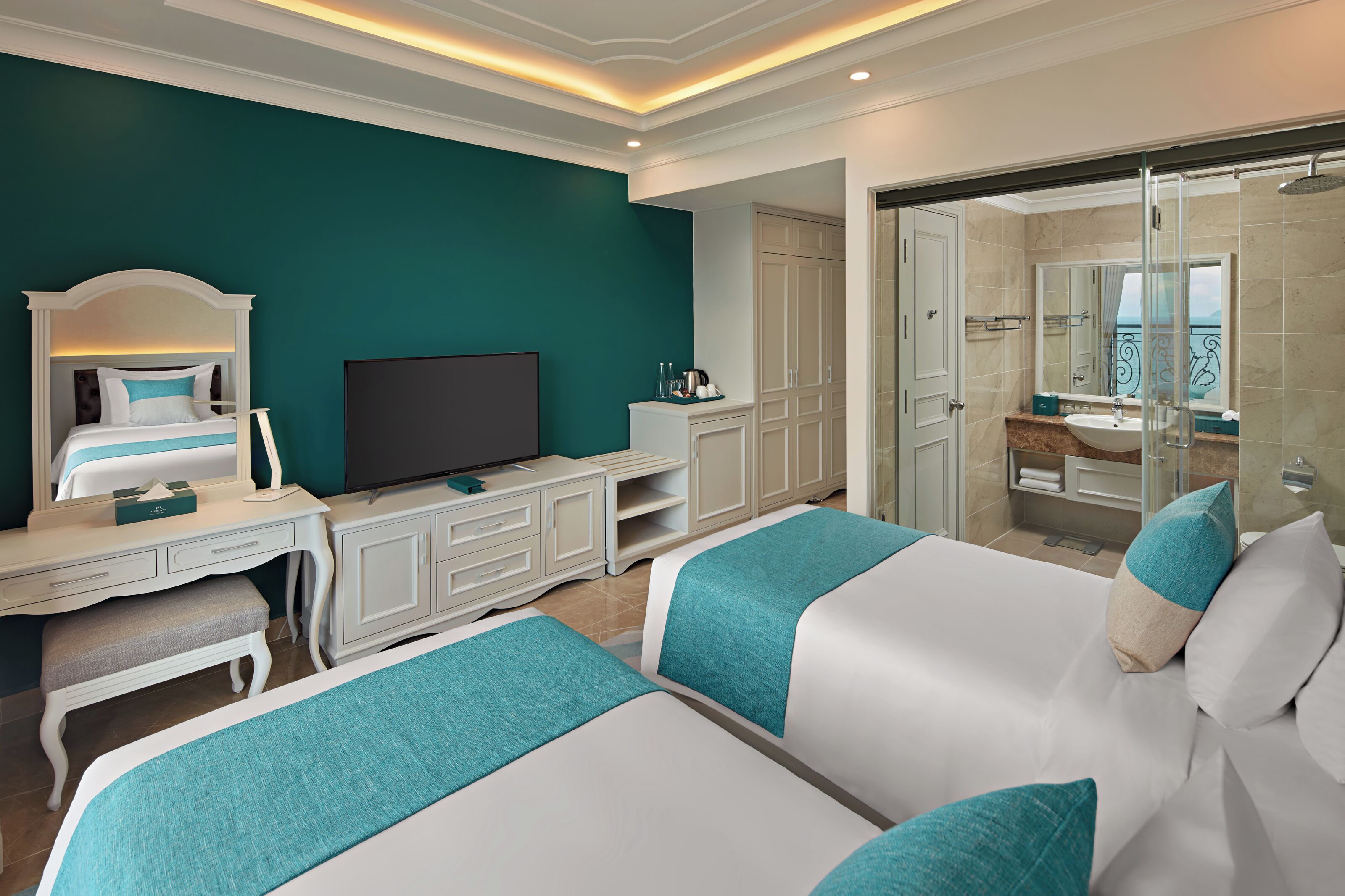 A hotel room with two beds, a TV, a desk, a wardrobe, and a bathroom with a glass shower.
