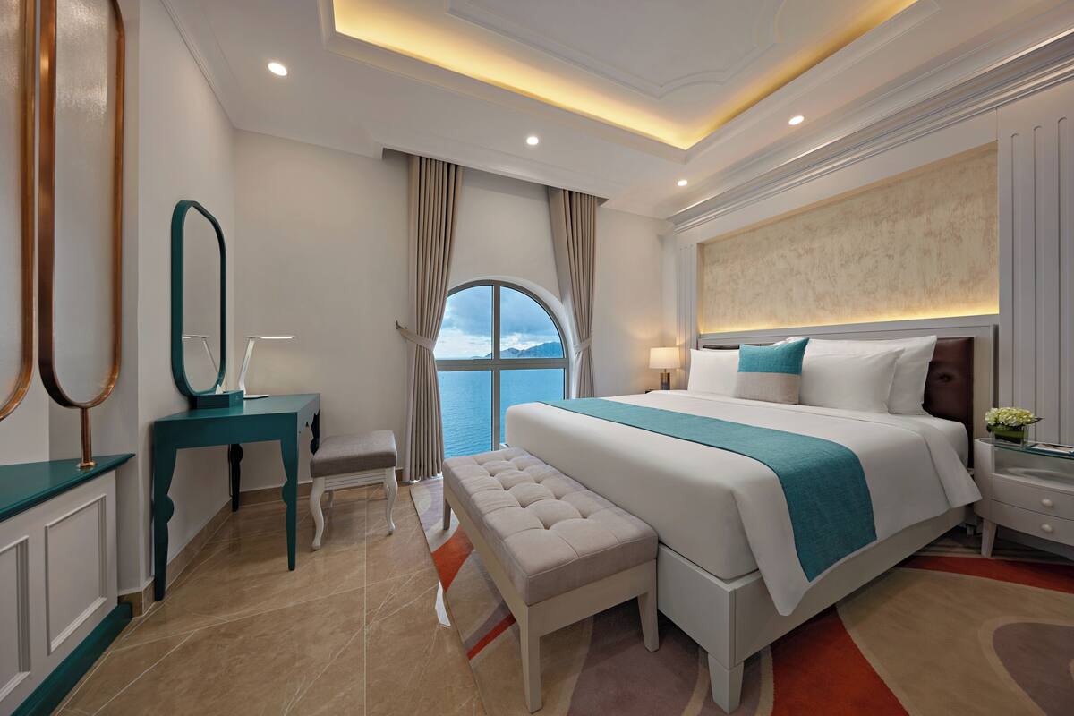 A modern hotel room with a large bed, a bench, a desk, and a view of the sea.
