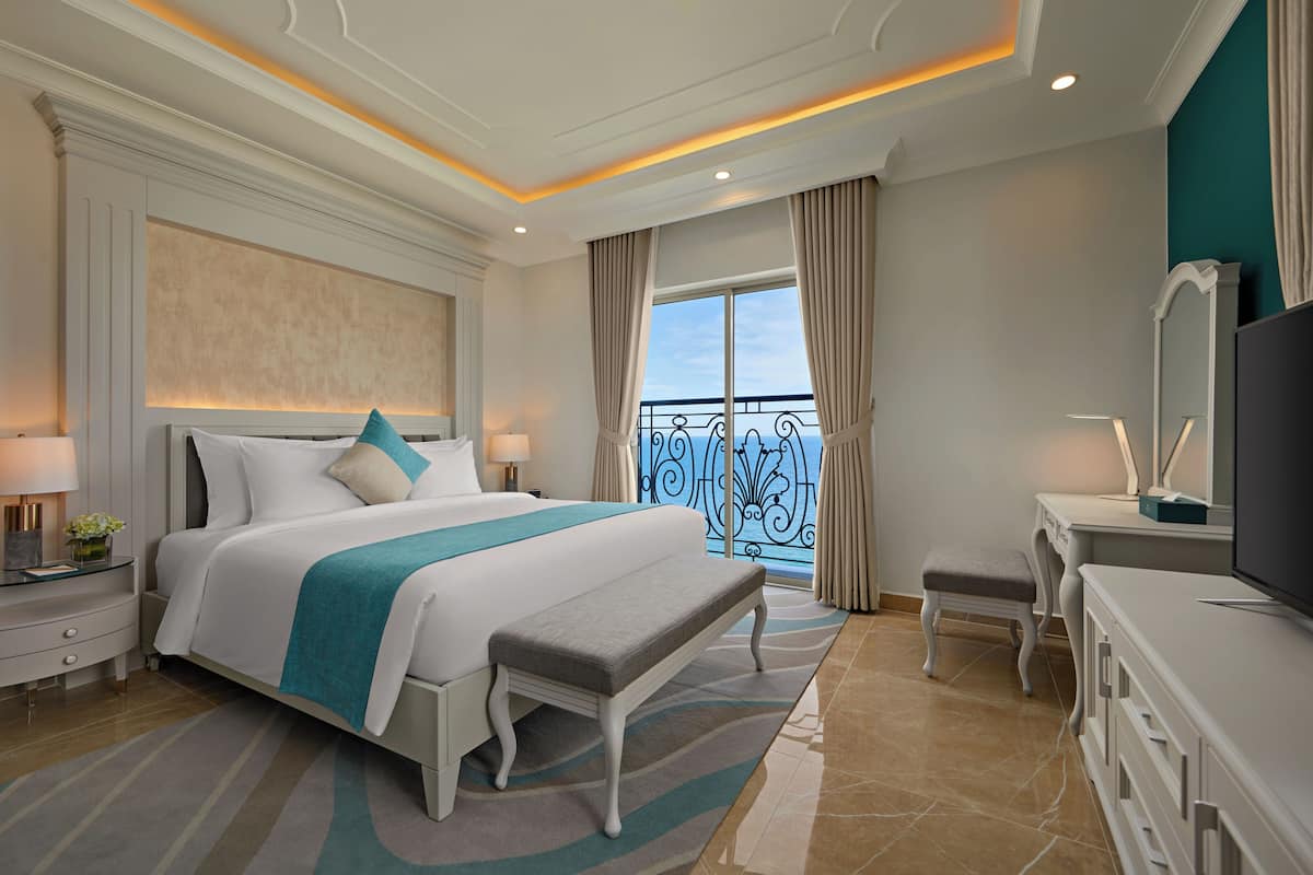 A hotel room with a large bed, a bench, a desk with a TV, and a balcony with a railing.