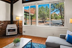 A living room with a fireplace, a sofa, a coffee table, and a large window with a view of red rock formations. - Charming Home w/Views! Susan Way - S052 (Sedona)