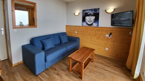 Ein Zimmer mit blauer Couch, einem Holztisch, einer Holzwandverkleidung, einem Wand-Fernseher und einem Fenster mit Holzrahmen.