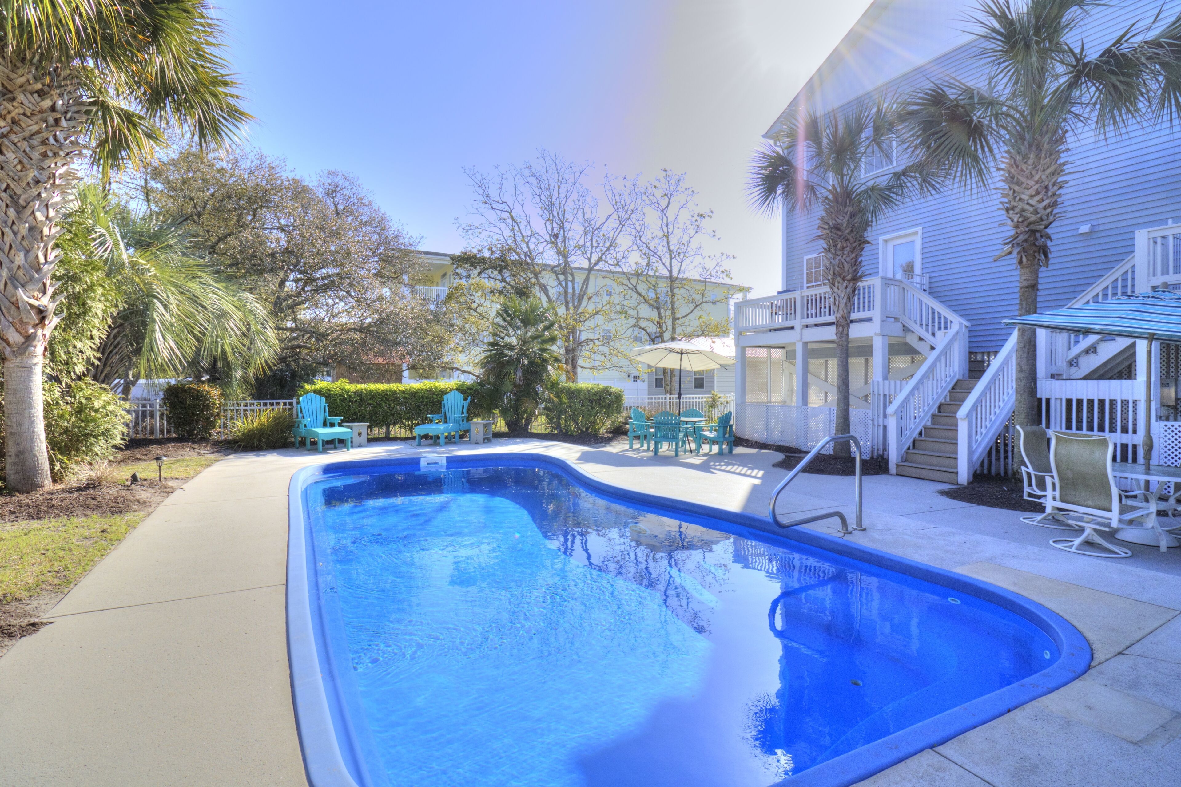 A swimming pool with a curved edge, surrounded by a concrete deck. There are chairs and a table in the background, and a building with a balcony and stairs nearby.