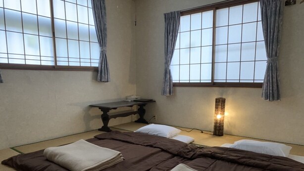 Une pièce de style japonais traditionnel, avec un sol recouvert de tatamis, une table basse et un lit doté d’une couverture de couleur marron foncé et d’oreillers blancs.