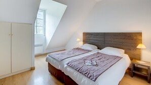 Un dormitorio con dos camas, cabecero de madera, una mesita de noche con lámpara y una ventana que deja entrar luz natural.