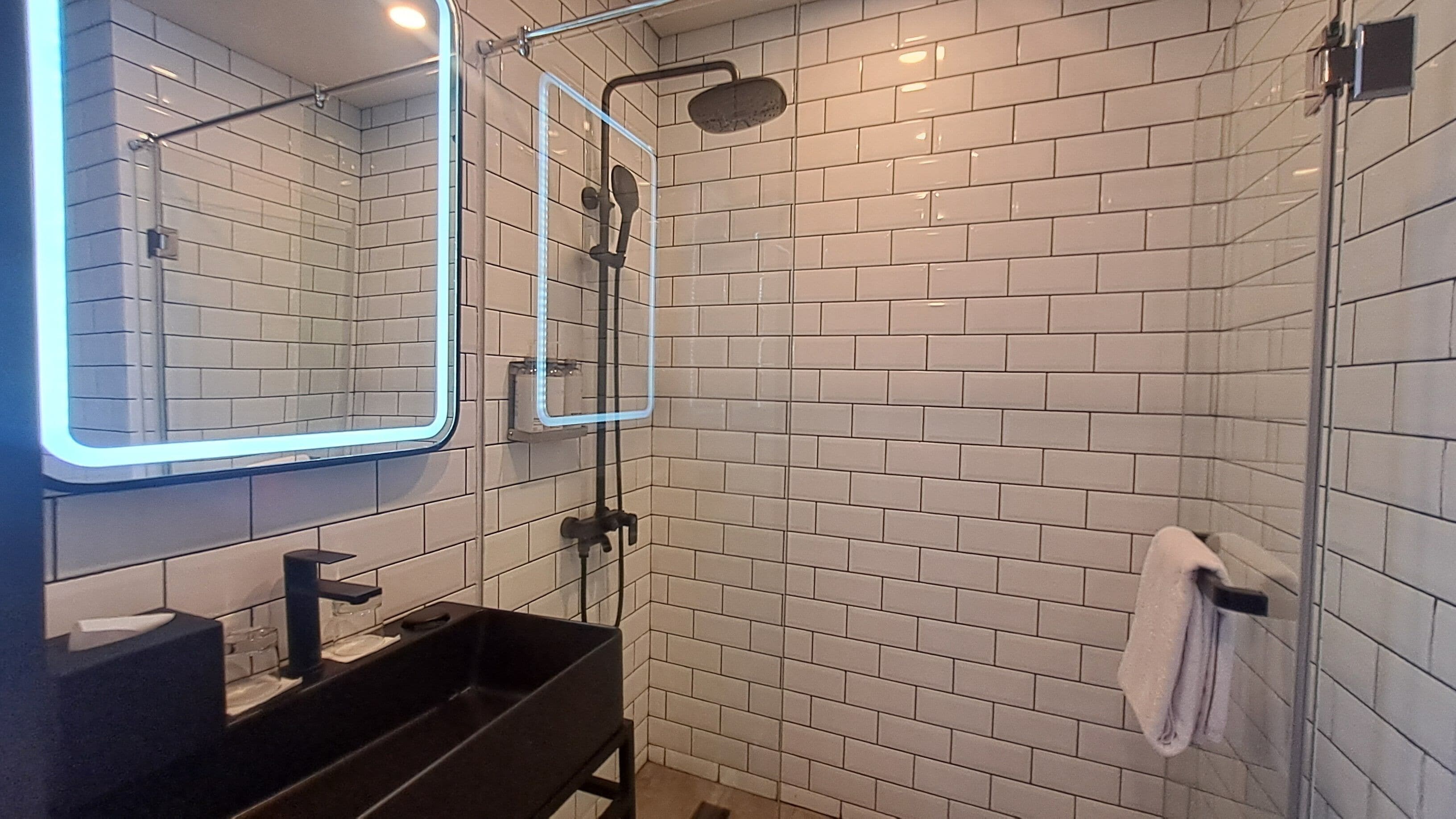 Un baño moderno con azulejos blancos tipo metro, un lavabo negro, un espejo y una ducha con cerramiento de vidrio.