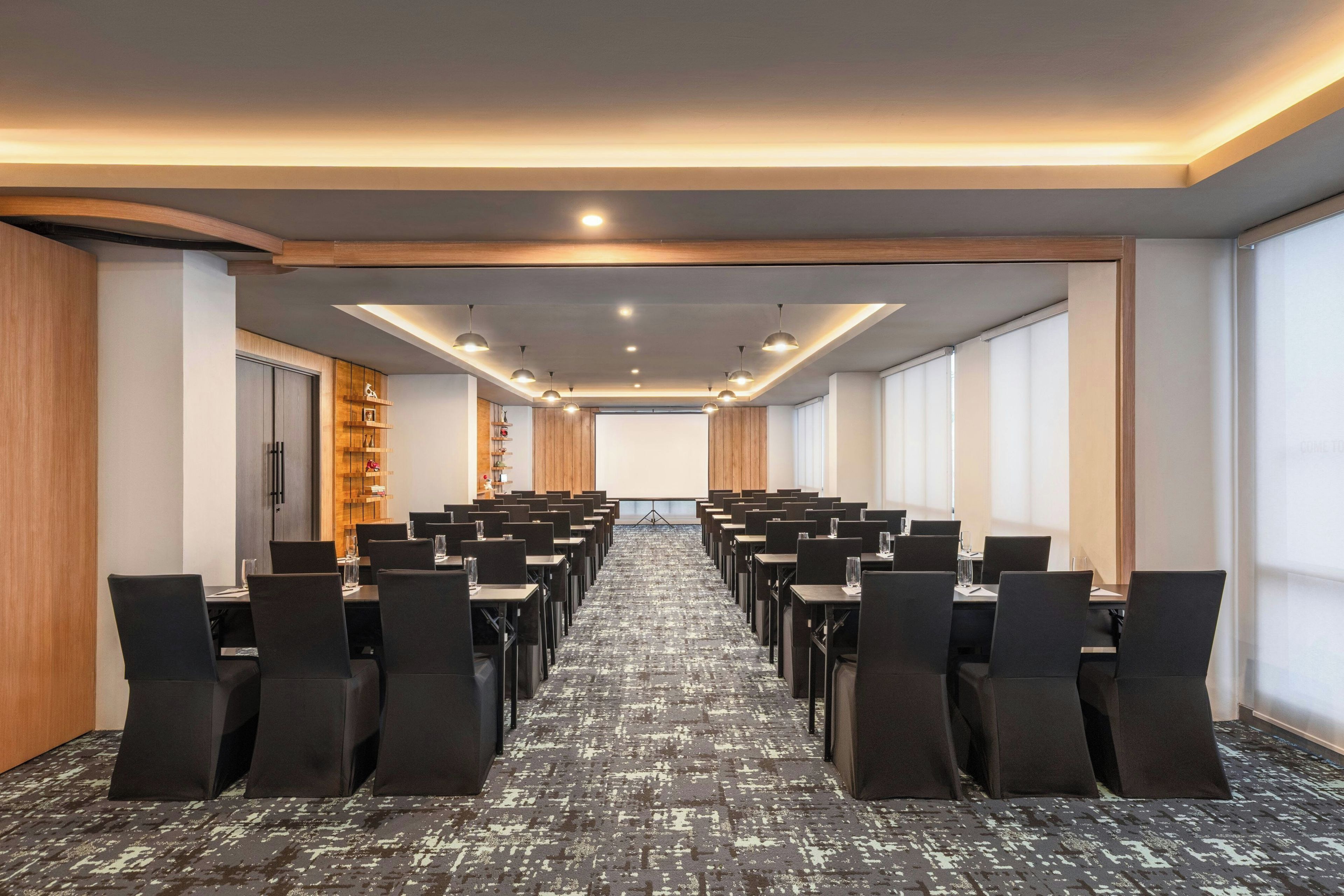 A conference room with rows of tables and chairs, a projector screen, and a wooden panel wall.