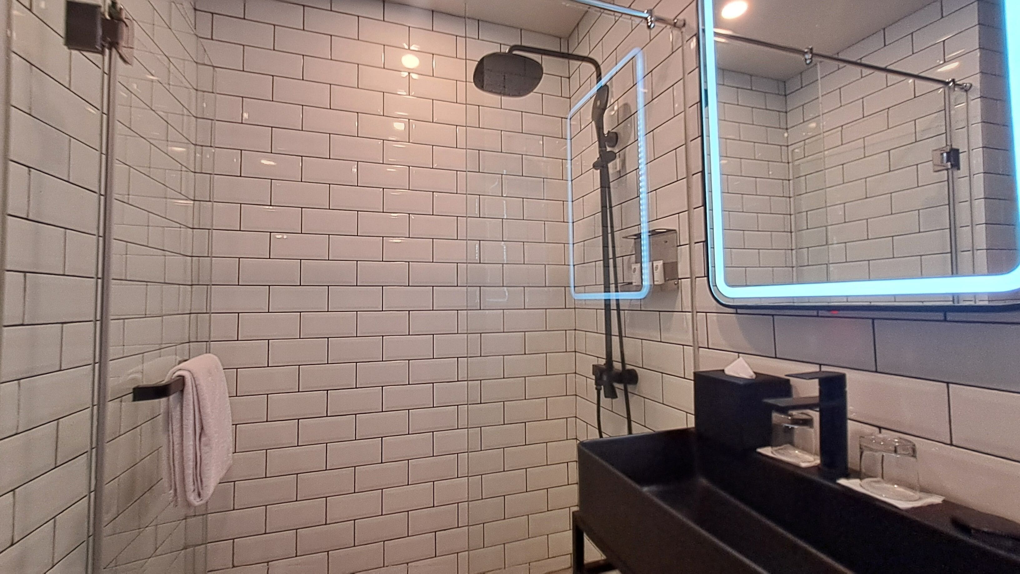 Un baño moderno con azulejos blancos tipo metro, ducha con cerramiento de vidrio, lavamanos negro y espejo con marco de neón.