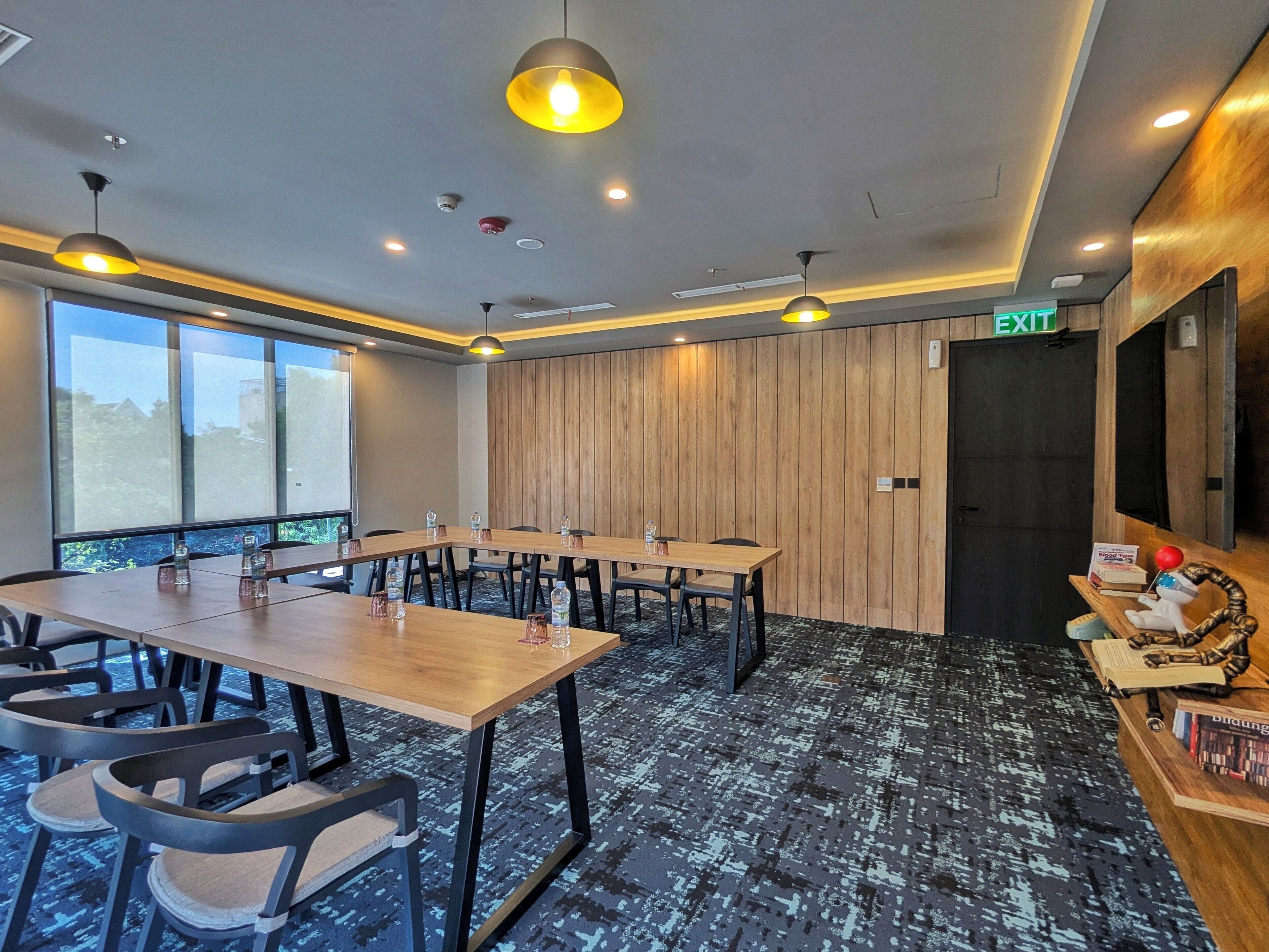 A conference room with a long table, chairs, and a wooden accent wall. There is an exit sign above a door, and a small shelf with books and a toy on the right.