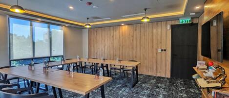 Un salón de conferencias con una larga mesa, sillas y una pared de acento de madera. Hay una señal de salida sobre una puerta, y una pequeña estantería con libros y un juguete a la derecha.