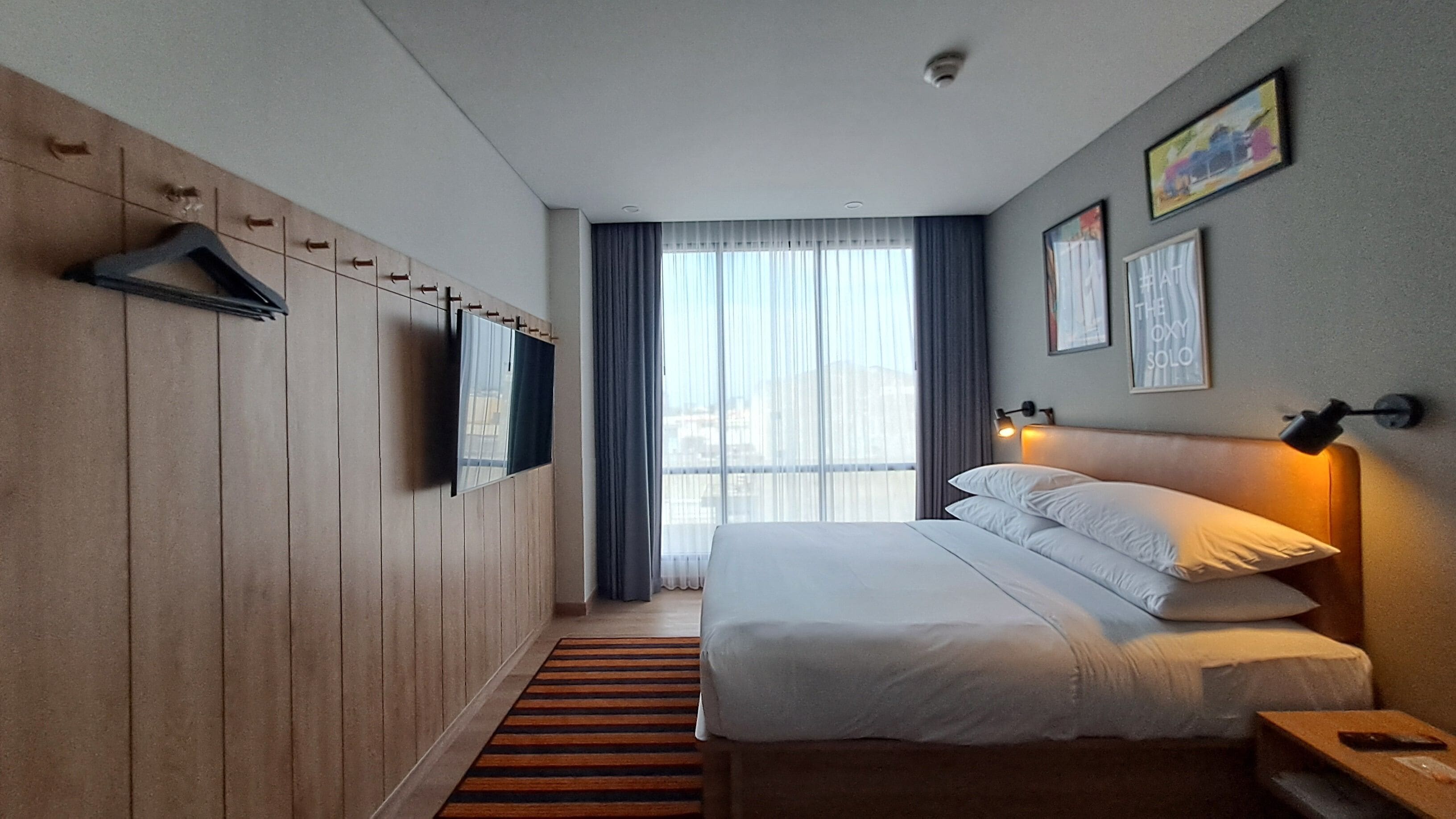 A hotel room with a bed, a TV mounted on the wall, a window with curtains, and a small table.