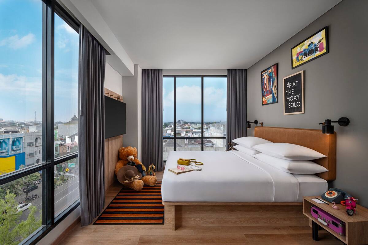 Una moderna camera d'albergo con un letto grande, un orsacchiotto, vista sulla città e opere d'arte alle pareti.