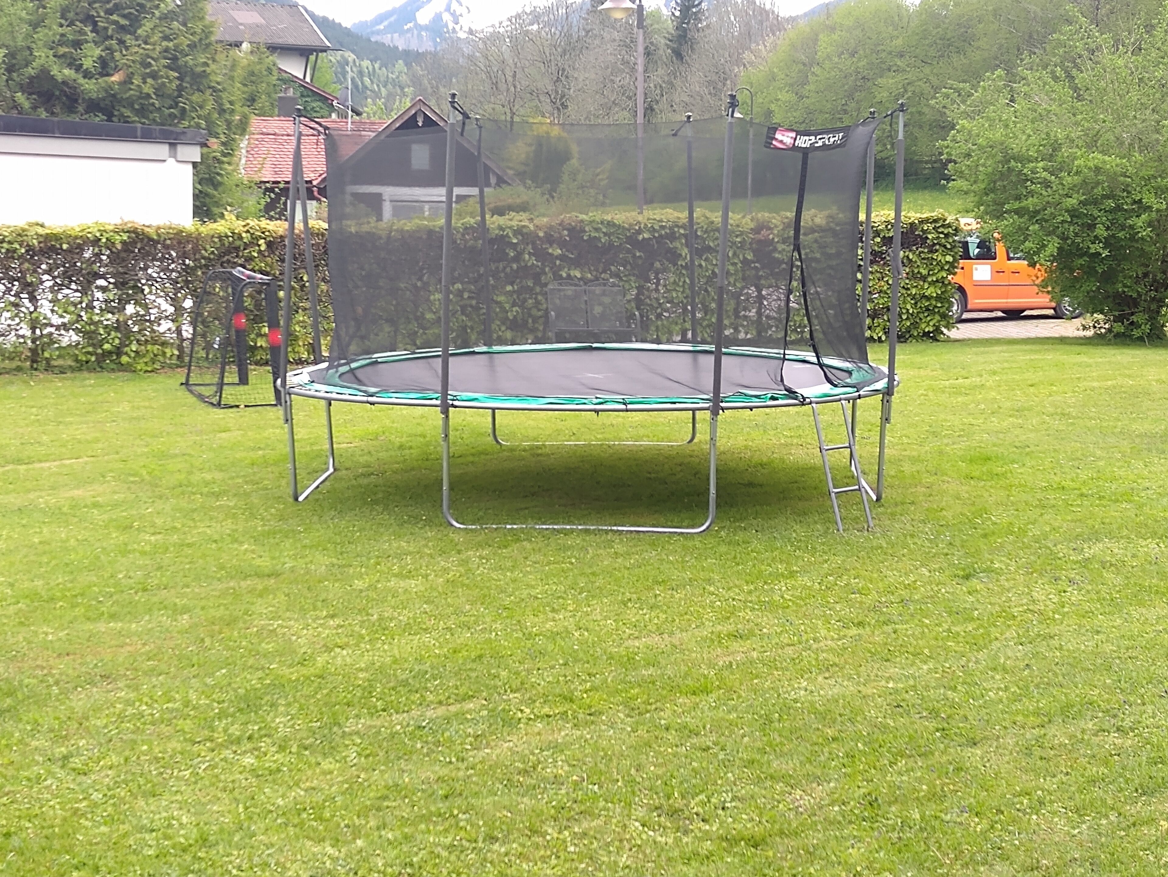 A trampoline in a grassy yard with a safety net, surrounded by trees and a house in the background.