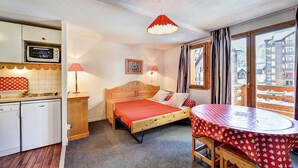 A cozy room with a bed, a small table, a kitchen area, and a window with a view of buildings. - Proche des Pistes + Téléski | Duplex avec Balcon (Risoul)