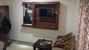 Une pièce avec un téléviseur fixé au mur, un mur en bois, une petite table avec une bouilloire et un plateau, et deux fauteuils en osier.