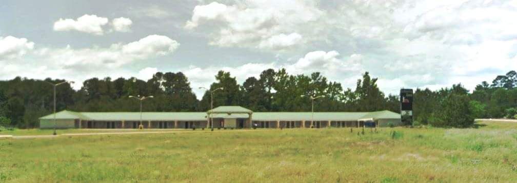 Un edificio grande y rectangular con techo plano, rodeado de un campo cubierto de hierba y árboles.