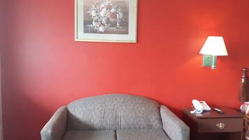 A room with a red wall, a framed floral artwork, a grey sofa, a bedside table with a lamp, and a telephone.