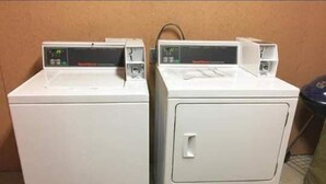Duas máquinas de lavar roupa brancas e de abertura frontal, lado a lado, numa divisão com uma parede lisa e um caixote de lixo no canto.