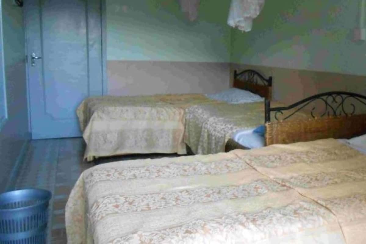 A room with two beds, each with a patterned bedspread and pillows. There is a blue trash bin and a door in the background.