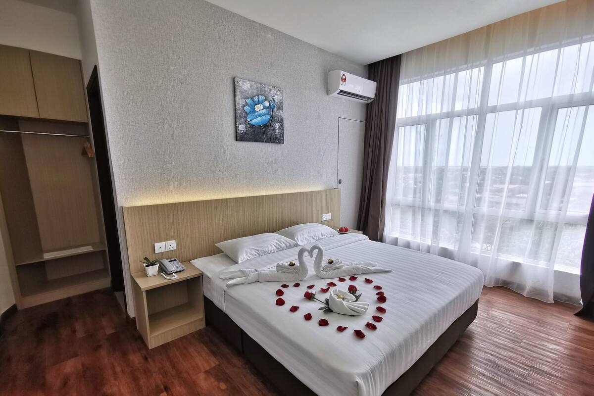 A hotel room with a large bed, a bedside phone, a painting on the wall, and a view through the window.