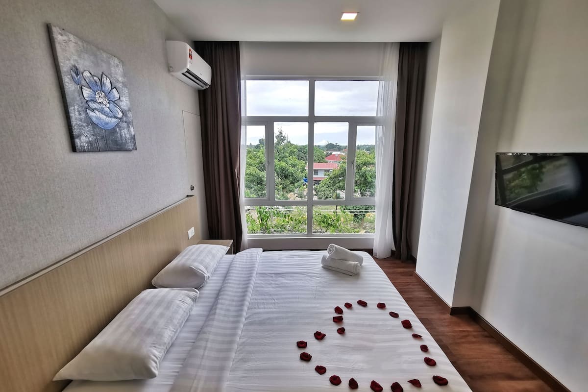 A hotel room with a bed, a painting on the wall, an air conditioning unit, a window with curtains, and a television mounted on the wall.