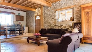 Un salon chaleureux avec un mur en pierre, des poutres apparentes en bois au plafond, un canapé, une table basse et un coin repas avec des chaises et une table.