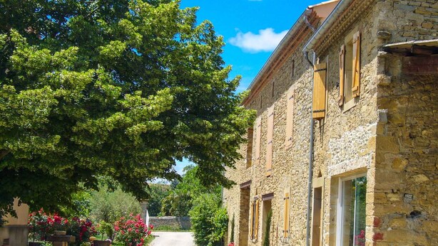 Un bâtiment en pierre avec une allée en gravier, entouré d’arbres et de plantes en pot.
