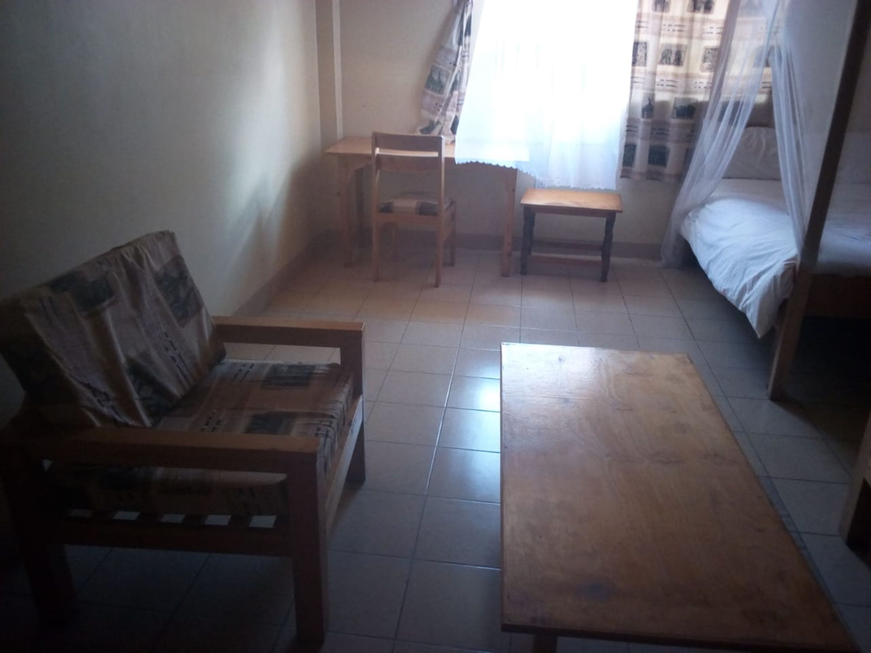 A small, dimly lit room with a single bed, a wooden bench, a small table, and a chair.