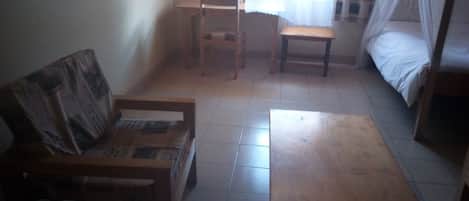 A small, dimly lit room with a single bed, a wooden bench, a small table, and a chair.