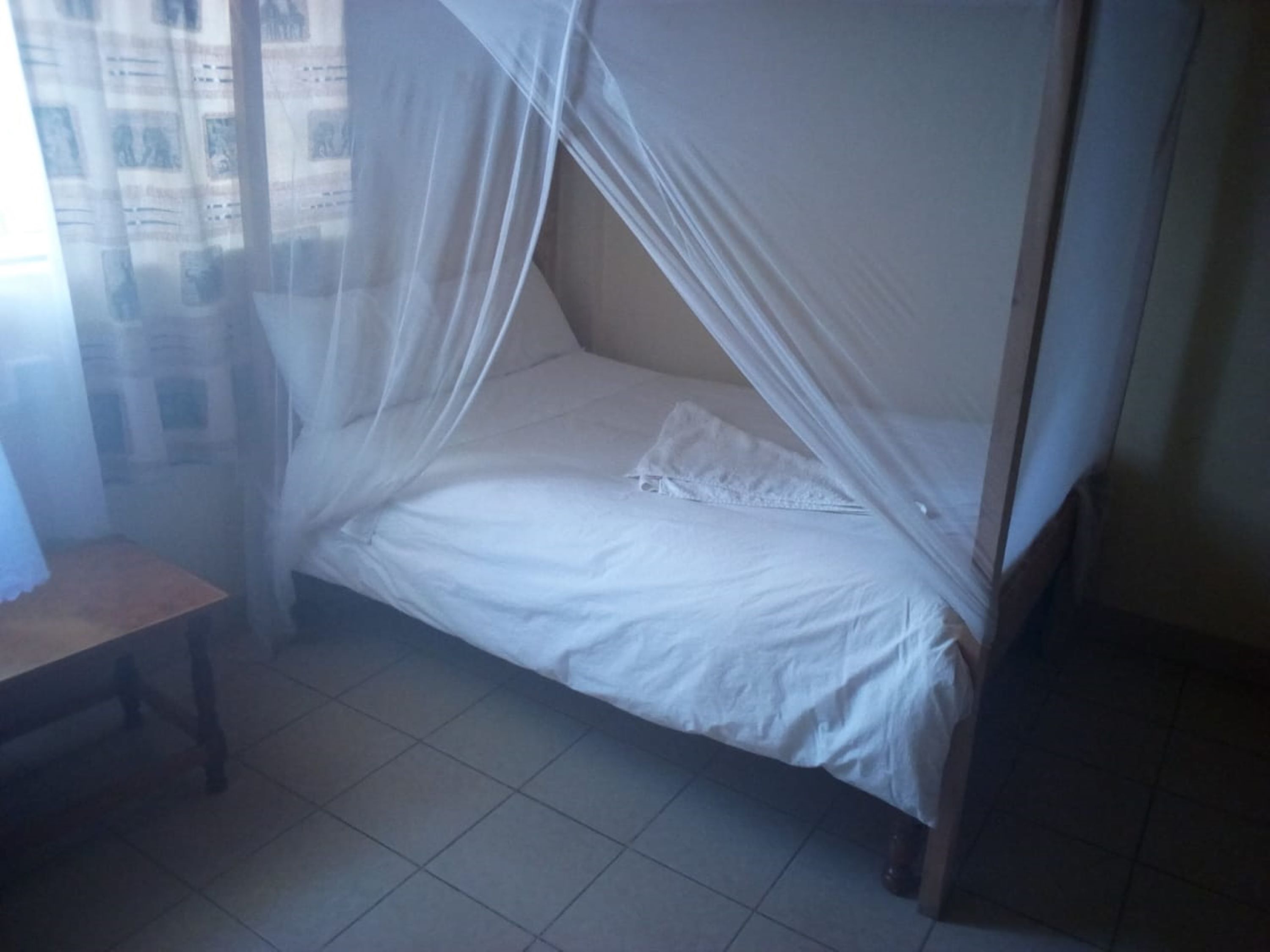 Uma cama de solteiro com mosquiteiro, uma mesinha de cabeceira de madeira e piso de azulejos.