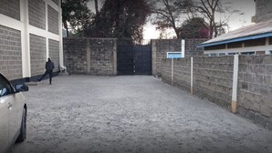 Uma área de estacionamento com um carro, uma pessoa caminhando e um prédio com parede de tijolos e telhado azul.