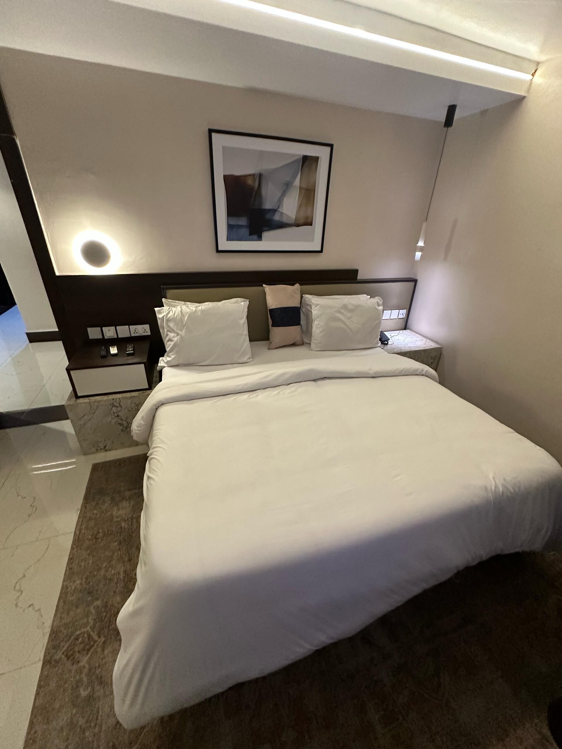 A modern hotel room with a large bed, a nightstand with a lamp, a framed artwork on the wall, and a marble floor.