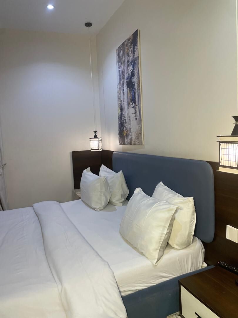 A neatly made bed with white bedding and pillows, a blue headboard, a bedside table with a lamp, and a large abstract painting on the wall.