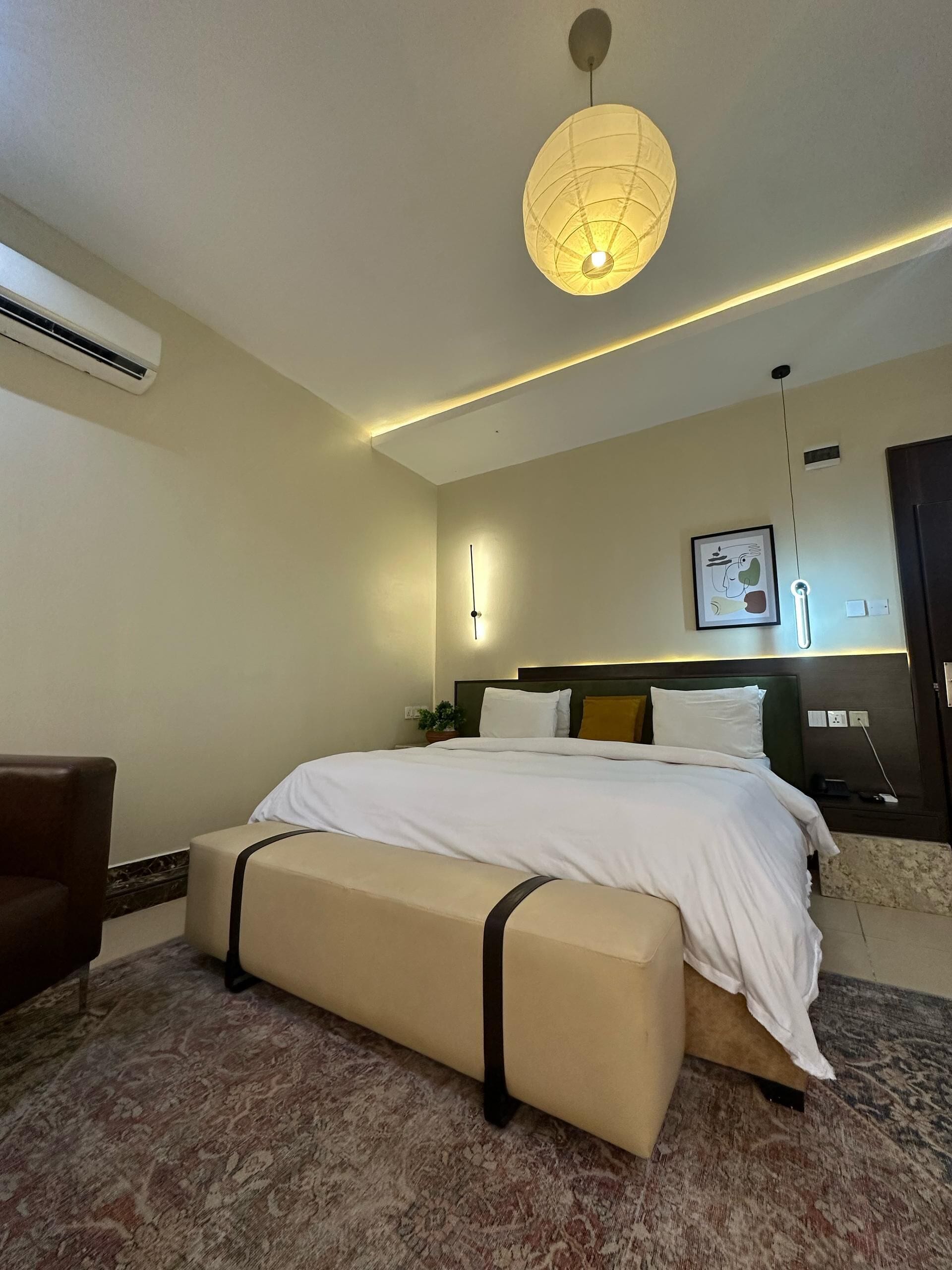 A hotel room with a bed, bedside table, lamp, air conditioning unit, and a patterned carpet.