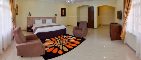 A spacious hotel room with a large bed, two armchairs, a desk, and a chandelier.