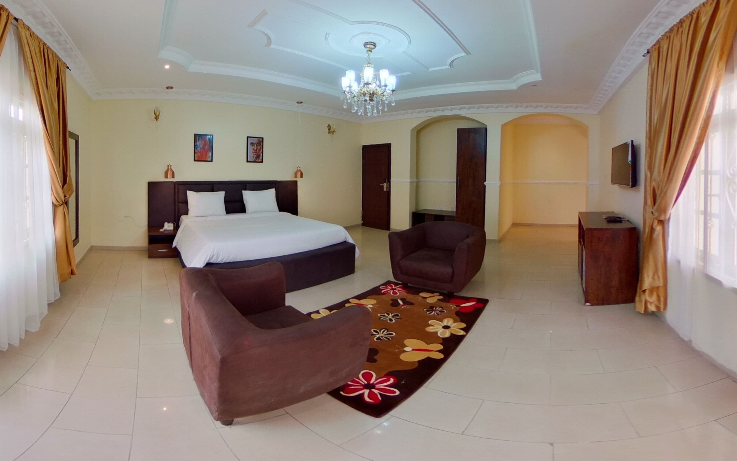 A spacious hotel room with a large bed, two armchairs, a TV, and a chandelier.