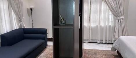 A modern hotel room with a sofa, a bed, a large rug, and a tall, dark-colored shelving unit.
